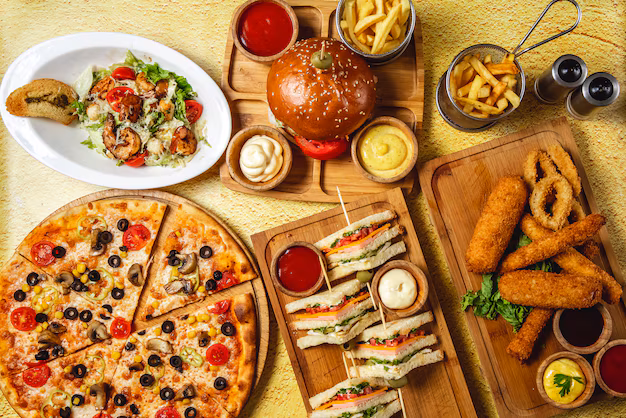 تصویر اصلی چیز فود، مجموعه‌ای از غذاهای فست فود شامل پیتزا، سوخاری و نوشیدنی روی میز چوبی با پس زمینه طلایی
